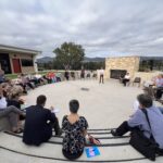 People sitting in an outdoor ampitheatre with trees and fields in the background and a brick fireplace in view