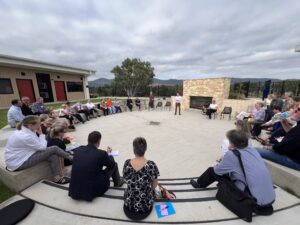 People sitting in an outdoor ampitheatre with trees and fields in the background and a brick fireplace in view